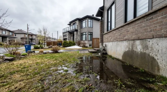Terrain résidentiel québécois avec flaque d'eau stagnante près des fondations après la fonte printanière, maison moderne en arrière-plan sous ciel couvert